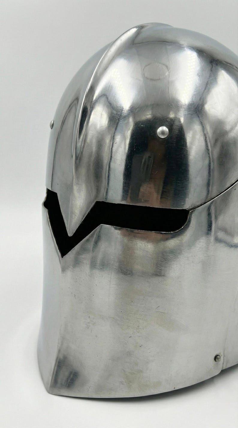 Puede incluir: Un casco plateado pulido con una distintiva abertura facial en forma de V. El casco, probablemente de metal, tiene una superficie lisa y reflectante y est&aacute; adornado con remaches. Su parte superior redondeada sugiere una est&eacute;tica medieval o fant&aacute;stica.