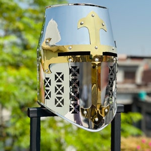 May include: A silver and gold knight's helmet with a face shield and a cross on top. The helmet is on a black stand.