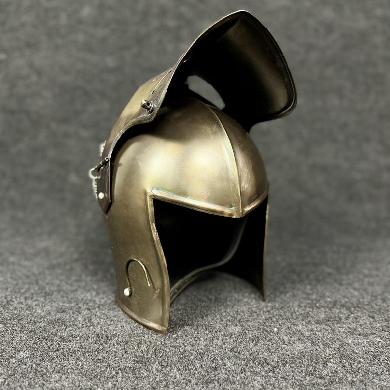 May include: A bronze-colored metal helmet with a wide, curved visor. The helmet has a rounded top and a face opening with a squared-off design. Decorative curved lines are on the sides. The helmet is likely for historical reenactment or display.