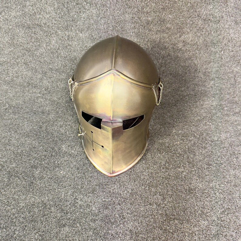 May include: A metallic silver knight's helmet with a visor featuring a cross design. The helmet has a rounded top and is detailed with small chains on each side. The surface has a slight iridescent sheen.