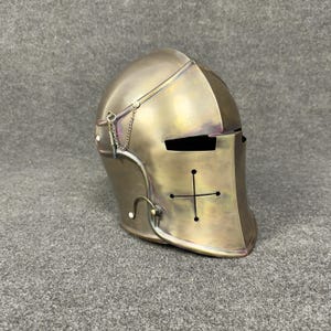 May include: A bronze-colored medieval helmet with a cross design on the front. The helmet features a visor with eye slits and a chain detail. The surface has a slightly aged appearance, suggesting historical use or a vintage aesthetic.
