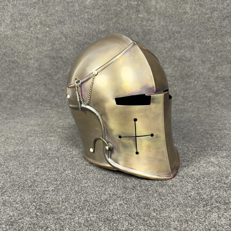 May include: A bronze-colored medieval helmet with a cross design on the front. The helmet features a visor with eye slits and a chain detail. The surface has a slightly aged appearance, suggesting historical use or a vintage aesthetic.