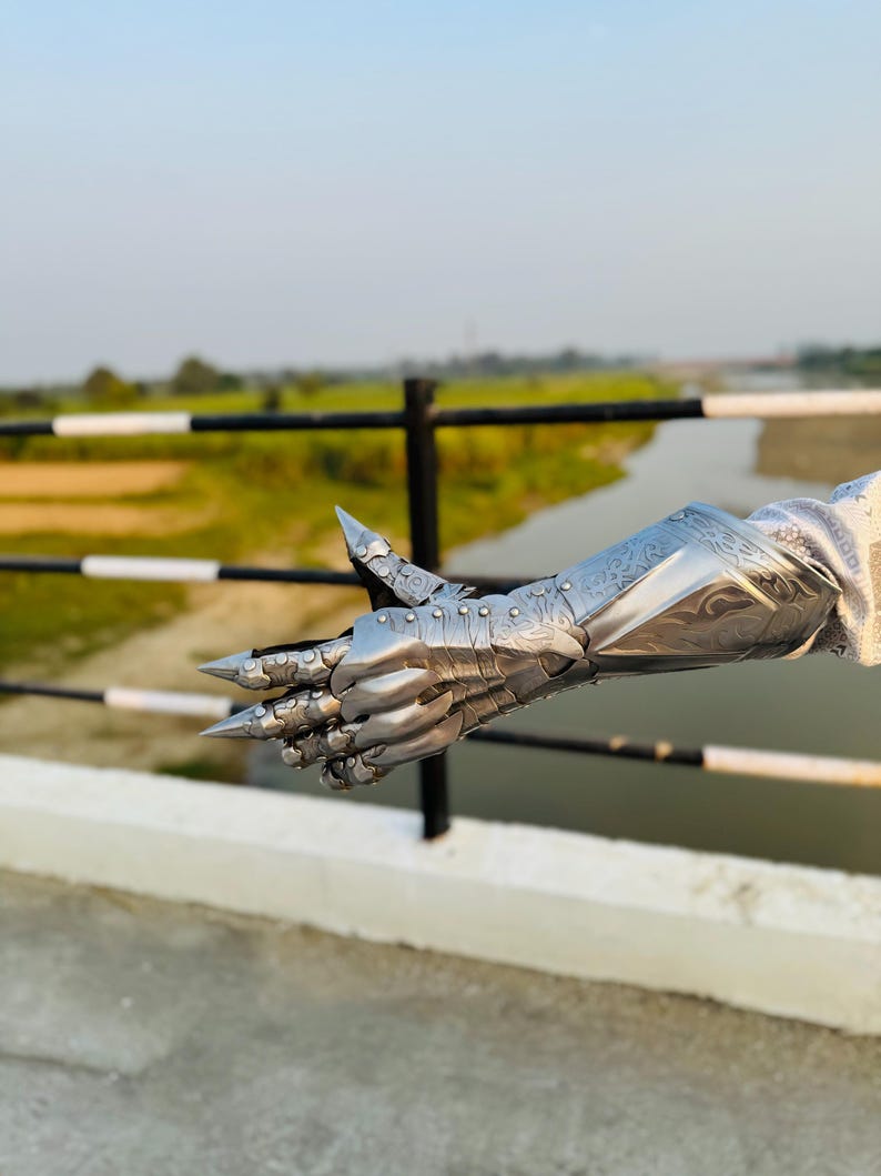 May include: A silver metal gauntlet with a claw-like design. The gauntlet is detailed with intricate engravings and rivets. The gauntlet is shown against a blurred background of a bridge and a river.