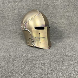 May include: A metallic gold-colored knight's helmet with a cross design on the front. The helmet features a visor and a chain detail on the side. The helmet is made of metal and has a vintage appearance.