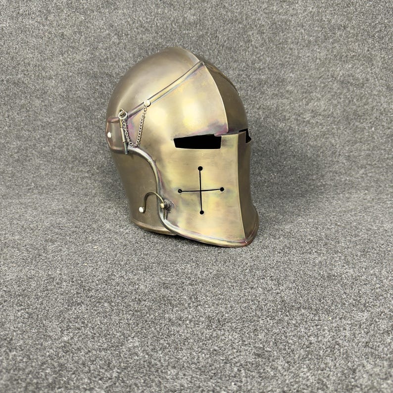 May include: A metallic gold-colored knight's helmet with a cross design on the front. The helmet features a visor and a chain detail on the side. The helmet is made of metal and has a vintage appearance.