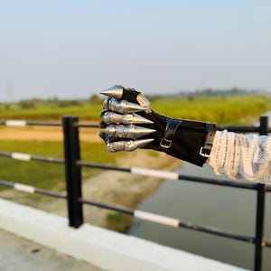 May include: A silver metal hand with pointed knuckles, worn as a glove, with a black leather wrist strap and buckle. The hand is clenched into a fist.