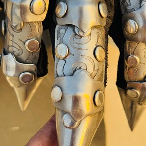 May include: Close-up of a silver metal glove with pointed finger tips. The glove has a decorative design and is made of metal plates.