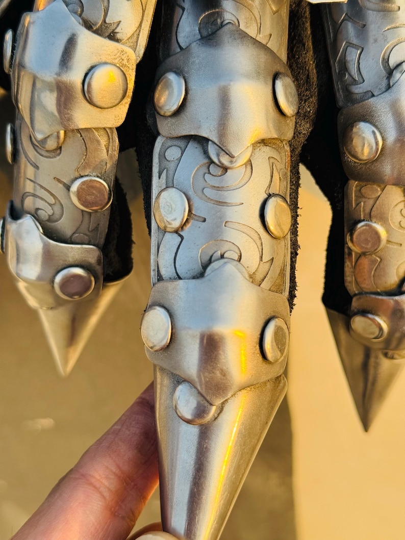 May include: Close-up of a silver metal glove with pointed finger tips. The glove has a decorative design and is made of metal plates.