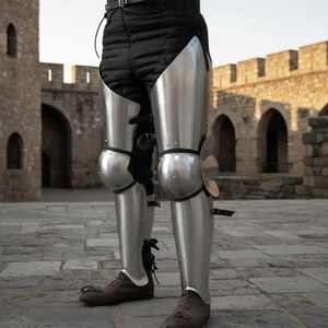 Puede incluir: Armadura de pierna de metal plateado, que incluye protecci&oacute;n para rodillas y espinillas, usada sobre pantalones acolchados negros. La armadura se asegura con correas y hebillas. El fondo presenta un edificio de piedra.