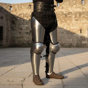 Puede incluir: Una armadura de piernas de acero pulido, con placas articuladas para los muslos y las rodillas. La armadura se lleva sobre pantalones acolchados negros y se asegura con un cintur&oacute;n de cuero. Las botas son de cuero marr&oacute;n. El fondo es una pared de piedra.
