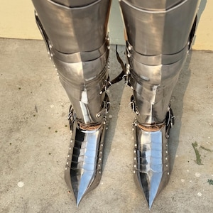 May include: A pair of silver-colored metal leg armor with pointed toes. The armor is made of multiple plates and is secured with straps and rivets.