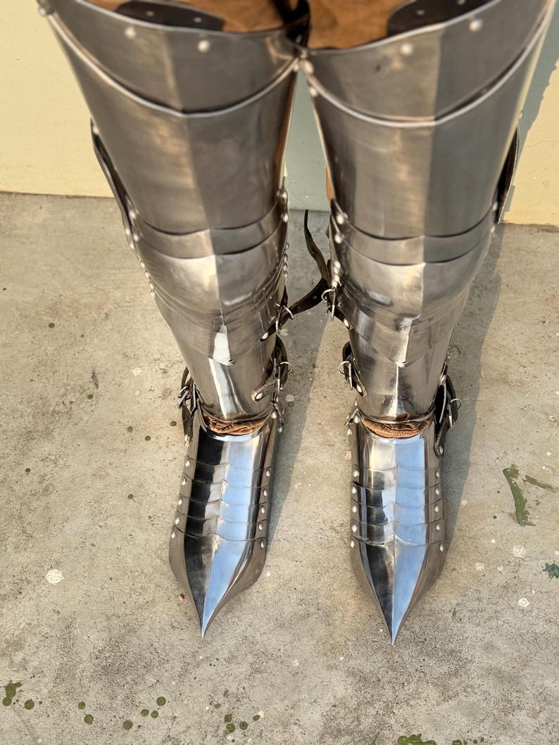 May include: A pair of silver-colored metal leg armor with pointed toes. The armor is made of multiple plates and is secured with straps and rivets.