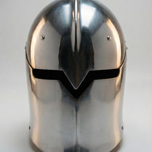 Puede incluir: Un yelmo medieval pulido, de color plateado, con una barbilla puntiaguda y una ranura horizontal para los ojos. El yelmo tiene una cresta central y est&aacute; remachado en los laterales. La superficie refleja la luz, creando reflejos y sombras.