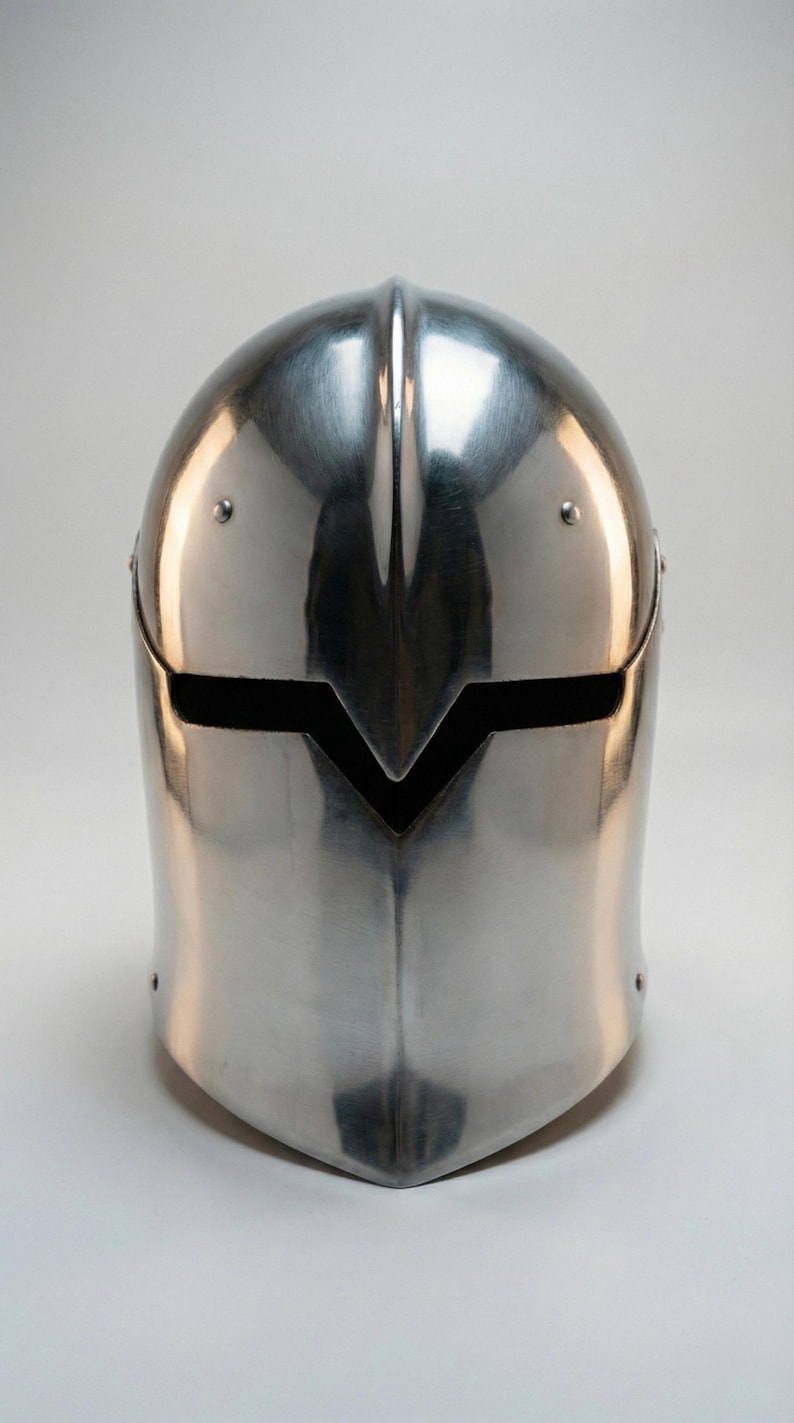 Puede incluir: Un yelmo medieval pulido, de color plateado, con una barbilla puntiaguda y una ranura horizontal para los ojos. El yelmo tiene una cresta central y est&aacute; remachado en los laterales. La superficie refleja la luz, creando reflejos y sombras.