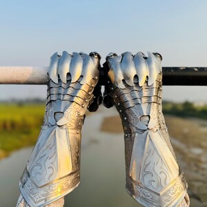 May include: A pair of silver metal gauntlets with intricate designs and pointed knuckles. The gauntlets are worn and have a rustic look.