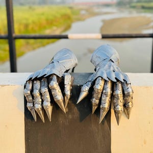 May include: A pair of silver metal gauntlets with pointed fingers and decorative details. The gauntlets are resting on a white surface.