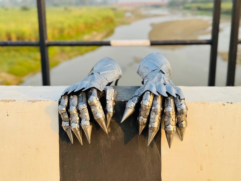 May include: A pair of silver metal gauntlets with pointed fingers and decorative details. The gauntlets are resting on a white surface.