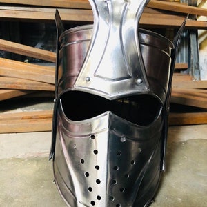 K&ouml;nnte beinhalten: Ein silberner Ritterhelm mit einer spitzen Spitze und einer Gesichtsmaske mit L&ouml;chern. Der Helm ist aus Metall und hat eine dunkle Oberfl&auml;che.