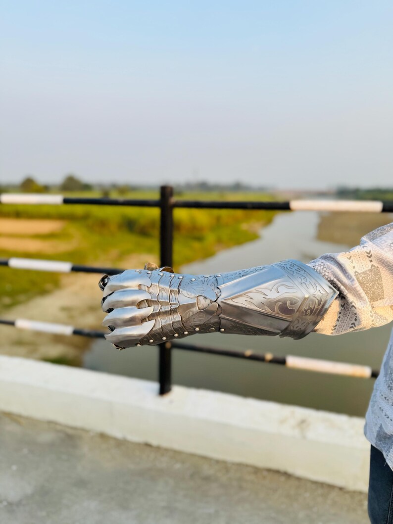 May include: A silver metal gauntlet with a decorative design. The gauntlet is worn on the left hand and extends to the elbow.