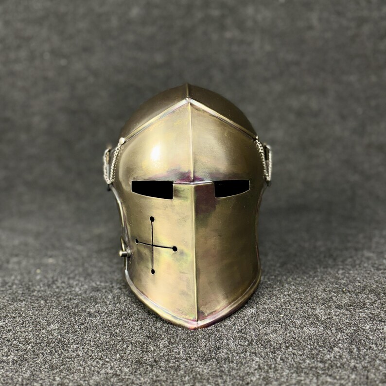 May include: A brass-colored medieval helmet with a cross design on the front. The helmet has eye slits and a chain detail on the sides. The metal has a polished finish, reflecting light.