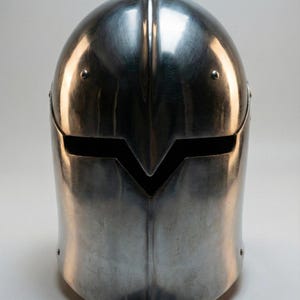 Puede incluir: Un casco pulido de color plateado con una distintiva abertura en forma de V en la cara. El casco tiene una superficie lisa y reflectante y est&aacute; dise&ntilde;ado con una parte superior redondeada y una parte inferior ligeramente puntiaguda. Se ven remaches.