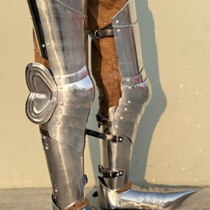 May include: A pair of silver metal shin guards with pointed toe boots. The shin guards are made of metal plates and have straps to secure them to the legs. The boots are also made of metal and have a pointed toe.