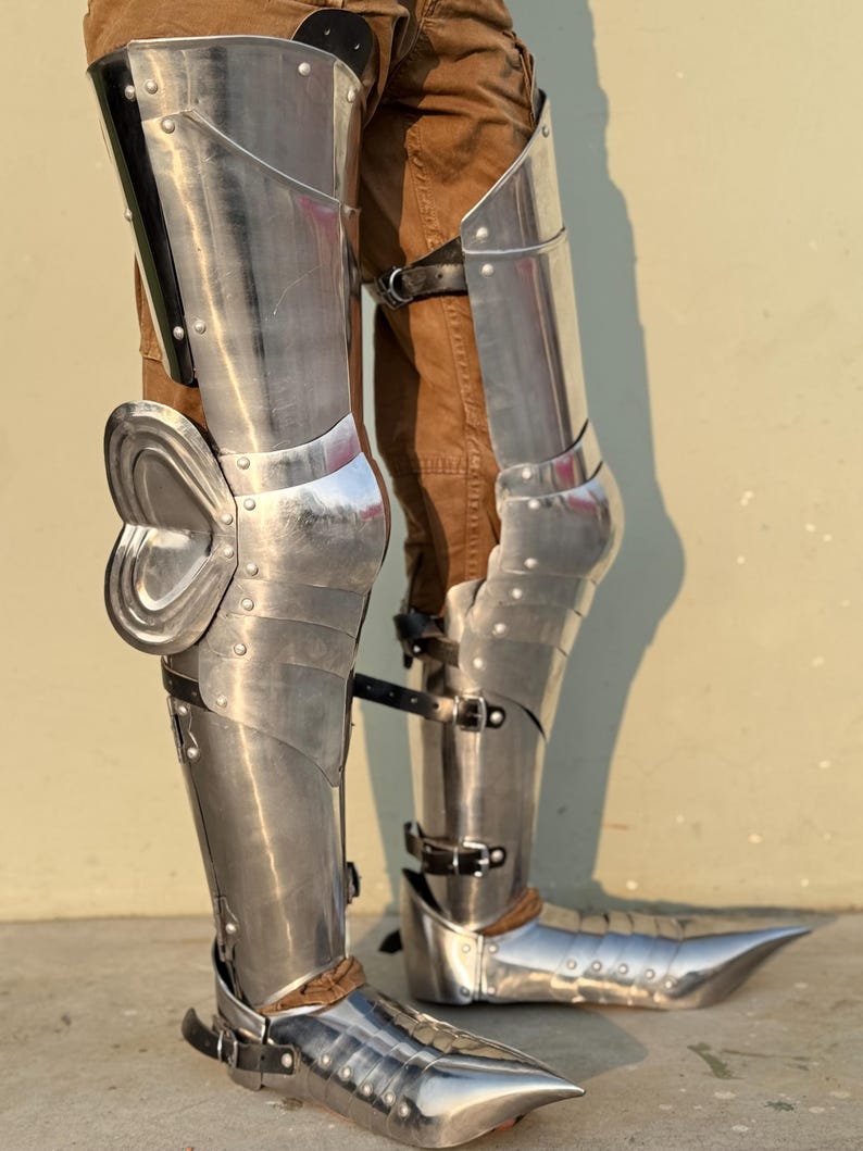 May include: A pair of silver metal shin guards with pointed toe boots. The shin guards are made of metal plates and have straps to secure them to the legs. The boots are also made of metal and have a pointed toe.
