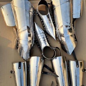 May include: A set of six silver metal shin guards with straps. The shin guards are made of metal plates and have a pointed design at the top. The straps are made of black leather.