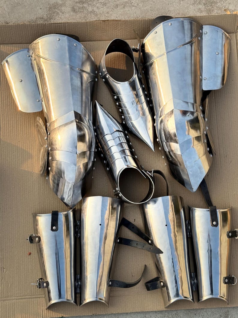 May include: A set of six silver metal shin guards with straps. The shin guards are made of metal plates and have a pointed design at the top. The straps are made of black leather.