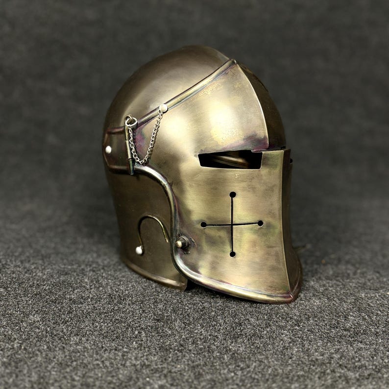May include: A bronze-colored medieval helmet with a cross design on the front. The helmet has a visor with eye slits and a chain detail. The surface shows signs of wear, giving it an antique appearance.