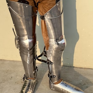 May include: A pair of silver metal leg armor with pointed toes. The armor is worn over brown pants.