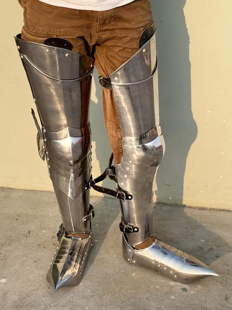 May include: A pair of silver metal leg armor with pointed toes. The armor is worn over brown pants.