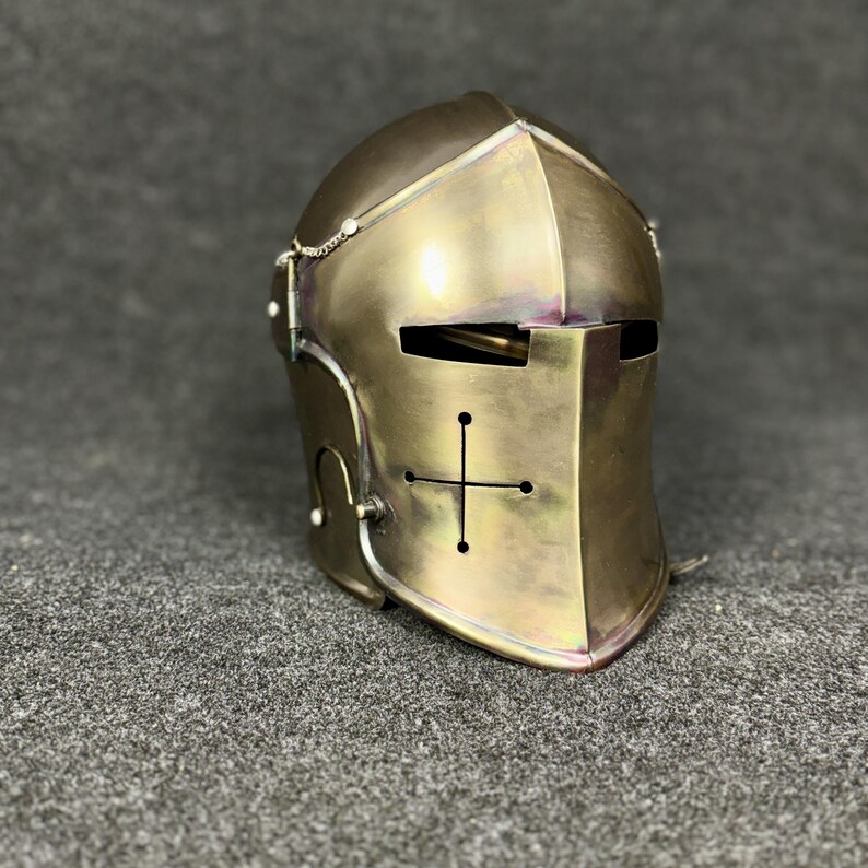 May include: A brass-colored medieval helmet with a cross design on the front. The helmet has a rounded top, a face guard with eye slits, and a chain detail. The surface has a polished finish, reflecting light.
