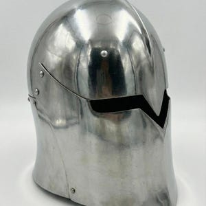 Puede incluir: Un casco de metal plateado pulido con una abertura para la visera. El casco tiene una parte superior redondeada y un borde inferior curvado. Los remaches aseguran los componentes del casco. La abertura de la visera tiene forma de V.