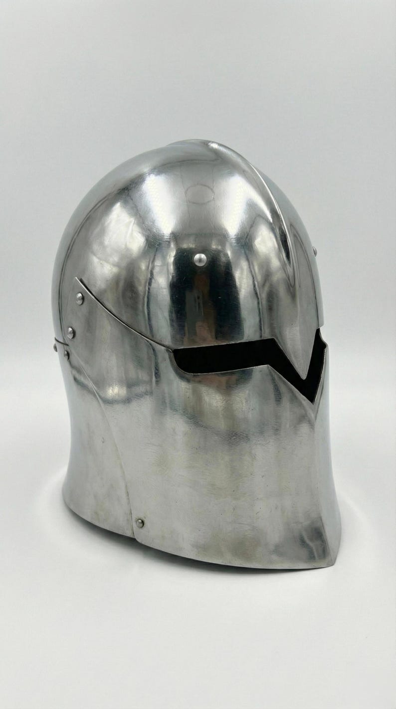 Puede incluir: Un casco de metal plateado pulido con una abertura para la visera. El casco tiene una parte superior redondeada y un borde inferior curvado. Los remaches aseguran los componentes del casco. La abertura de la visera tiene forma de V.