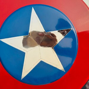 Close-up view of a Captain America-style shield. The shield is predominantly red and blue with a large metallic silver star at its center. The star has a five pointed design and is sharply defined against the blue background. The red outer ring of the shield is wide and forms a bold contrast to the blue inner circle. The shield's surface appears smooth and glossy, suggesting a polished finish. The colors are vibrant and saturated, the overall impression is one of a high-quality collectible item.