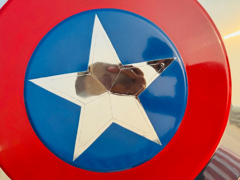 Close-up view of a Captain America-style shield. The shield is predominantly red and blue with a large metallic silver star at its center. The star has a five pointed design and is sharply defined against the blue background. The red outer ring of the shield is wide and forms a bold contrast to the blue inner circle. The shield's surface appears smooth and glossy, suggesting a polished finish. The colors are vibrant and saturated, the overall impression is one of a high-quality collectible item.