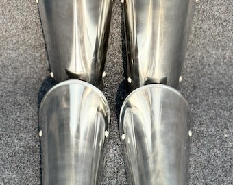 May include: A pair of silver shin guards with a rounded, slightly flared shape. The guards are made of metal and have a smooth, polished finish. They are secured with small rivets.