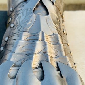 May include: Close-up of a silver metal gauntlet with intricate designs and a pointed finger guard. The gauntlet is made of multiple plates that are joined together with rivets.