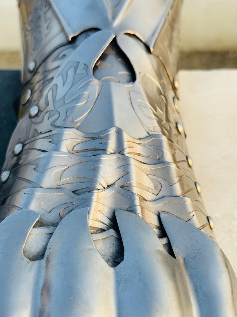 May include: Close-up of a silver metal gauntlet with intricate designs and a pointed finger guard. The gauntlet is made of multiple plates that are joined together with rivets.