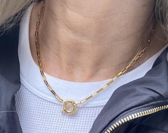 May include: A gold chain necklace with a round clasp. The necklace is worn by a person wearing a white shirt and a black jacket.