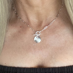 May include: A silver-toned necklace featuring a chain with elongated oval links and a toggle clasp closure. A small, circular, reflective silver-toned pendant hangs from the centre of the toggle mechanism, resting against the chest area.