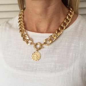 May include: A gold chain necklace with a large, chunky link design. The necklace has a round, gold pendant with a detailed design.
