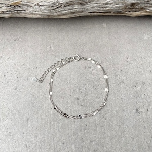 Könnte beinhalten: Ein silbernes Kettenarmband mit einem zarten, geschichteten Design. Das Armband besteht aus zwei Ketten, eine mit kleinen, quadratischen Gliedern und die andere mit einem traditionelleren Kettenglied-Stil.