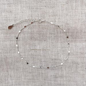 Peut inclure: Une fine chevillère en chaîne argentée ornée de petits éléments métalliques plats et réfléchissants répartis uniformément. Le bijou comprend une chaînette d'extension fine terminée par une breloque en forme de goutte et un fermoir à ressort.