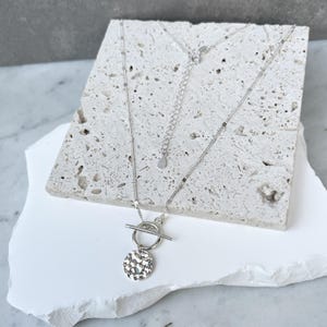 May include: A silver necklace featuring a hammered disc pendant and a toggle clasp. The necklace is presented on a white, textured surface. The chain is delicate and the pendant has a textured finish.