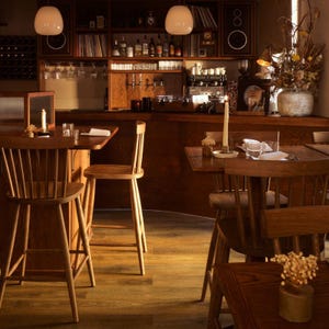 K&ouml;nnte beinhalten: Eine Holzbar mit einer geschwungenen Theke und einem Holzregal mit Flaschen und Gl&auml;sern. Es gibt zwei Holzst&uuml;hle an der Bar und zwei Holzst&uuml;hle an einem Tisch mit einer wei&szlig;en Tischdecke. Die Bar wird von zwei Pendelleuchten beleuchtet.