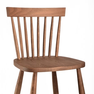K&ouml;nnte beinhalten: Ein brauner Holzstuhl mit einer Lattenlehne und einer abgerundeten Sitzfl&auml;che. Der Stuhl hat ein schlichtes, klassisches Design.