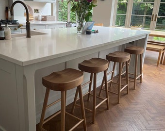 May include: Four wooden bar stools with a curved seat and a light brown finish. The stools are arranged in a row in front of a white kitchen island with a white countertop. The floor is a dark brown herringbone pattern.