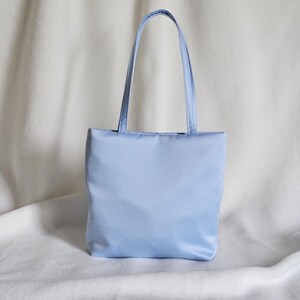 May include: A light blue tote bag with a smooth, solid-colored fabric exterior. The bag features a rectangular body and two matching light blue shoulder straps attached to the top edge, providing a simple and functional design for carrying items.